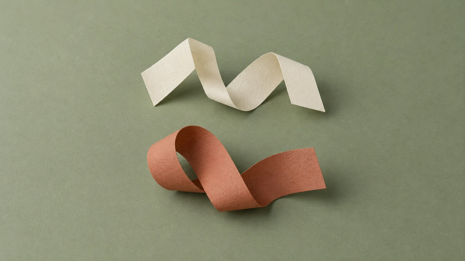 Two distinct folded-paper sculptures, one ivory and one terracotta, rest separately on a sage surface.